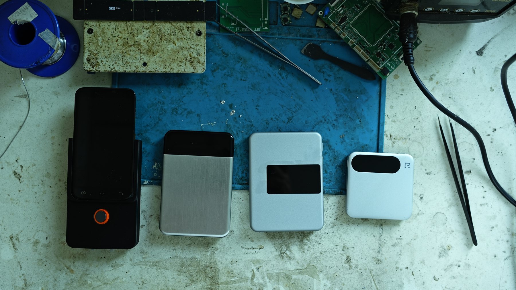 Four generations of the NoteCapt device lined up on a workbench, next to a soldering mat, tweezers and a bare circuit board
