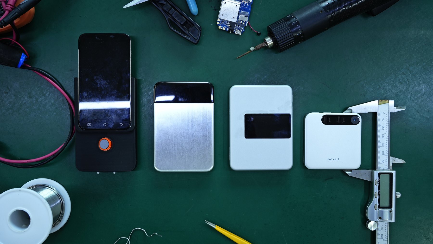 The same four generations on a green workbench, with a digital caliper beside the current model for scale